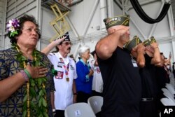 Personas que asistieron a una ceremonia por la llegada de los restos de se supone pertenecen a soldados que murieron en la Guerra de Corea, se ponen de pie durante el Himno Nacional en la Base Conjunta Pearl Harbor-Hickam, Hawai, el miércoles 1 de agosto de 2018.