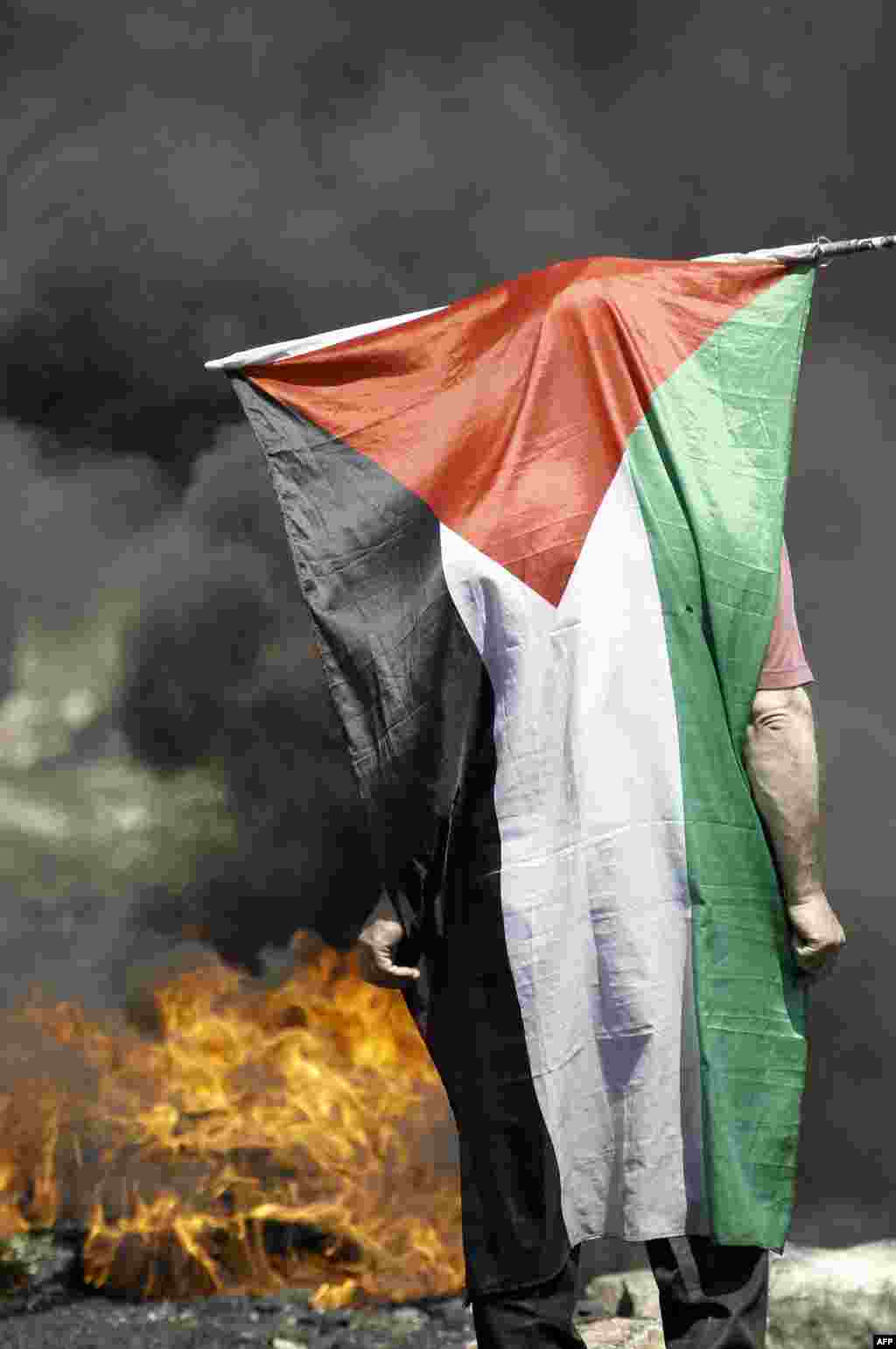 A Palestinian protestor stands with his national flag draped over himself near burning tires during clashes with Israeli soldiers following a demonstration against the expropriation of Palestinian land by Israel in the occupied West Bank.