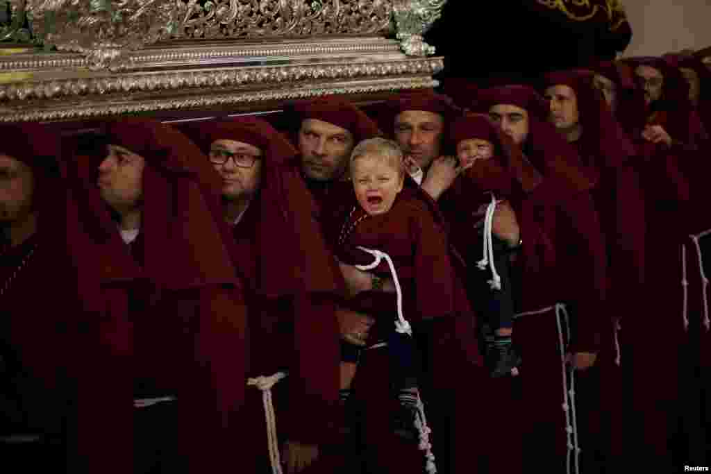 The 18-month-old fraternal twins, Nacho Guirado (L) and Asier cry as they are carried by their father Ignacio (L) and their uncle Jose Carlos, who are twins, while lifting a float bearing the statue of the Virgin inside a church as they take part as penitents in the &quot;Salud&quot; brotherhood in a Palm Sunday procession at the start of Holy Week in Malaga, southern Spain, March 29, 2015.