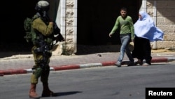 Palestinians walk near an Israeli soldier as he stands guard during an operation to locate three Israeli teens in the West Bank City of Hebron, June 17, 2014.
