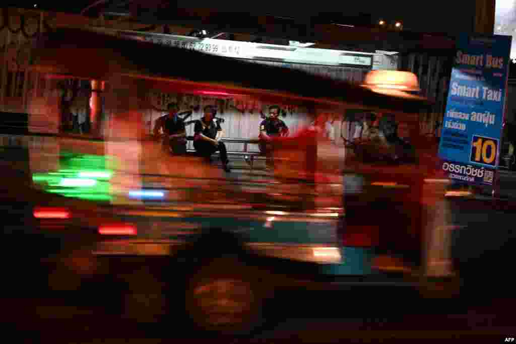 A tuktuk drives past people waiting for buses at a bus stop in Bangkok, Thailand.