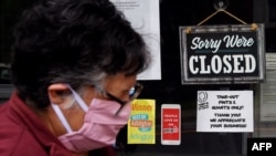 (FILES) In this file photo taken on May 4, 2020, a woman wearing a face mask walks past a sign in the window of a food store announcing that the business is closed during a shelter in place lockdown order during an outbreak of COVID-1 in Arlington, Virgin