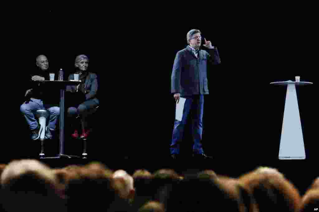 People sit at a table on stage and look towards the hologram of hard-left French presidential candidate Jean-Luc Melenchon, as he speaks to supporters who are gathered in Saint-Denis, near Paris.