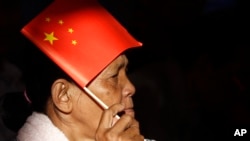 In this July 2, 2018 photo, a woman uses a Chinese national flag to protect herself from the sun as she listens to PM Hun Sen who delivering his speech during an inauguration ceremony of a sky bridge in Phnom Penh, Cambodia.