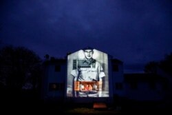 An image of veteran Charles Lowell is projected onto the home he shared with his wife, Alice, for 30 years as she stands at left with her daughter, Susan Kenney, in Hardwick, Mass., May 2, 2020.