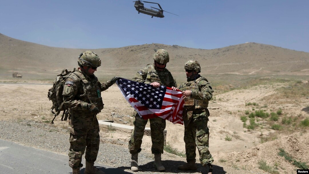 Tentara NATO berpose sambil memegang bendera Amerika sementara helikopter Chinook lepas landar seusai serah terima pengalihan tanggung jawab keamanan di akademi militer di luar Kabul (18/6). Afghanistan menangguhkan perundingan bilateral dengan AS, menyusul rencana dialog AS-Taliban.