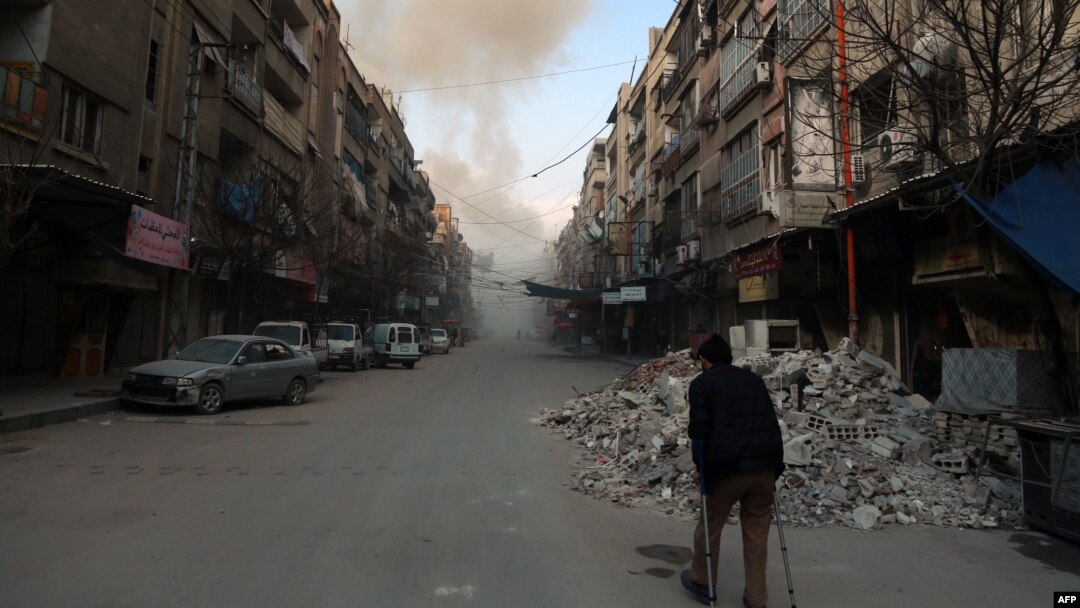 FILE - A Syrian man on crutches walks down a street as smoke billows in the rebel-held town of Douma, in the besieged Eastern Ghouta region on the outskirts of the capital Damascus, following air strikes by regime forces on the area on Feb. 23, 2018.