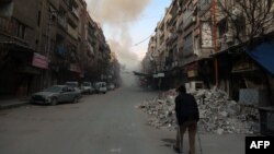 FILE - A Syrian man on crutches walks down a street as smoke billows in the rebel-held town of Douma, in the besieged Eastern Ghouta region on the outskirts of the capital Damascus, following air strikes by regime forces on the area on Feb. 23, 2018.