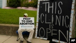 FILE - An abortion opponent sits behind a sign that advises the Women's Health Organization clinic is still open in Jackson, Mississippi, July 6, 2022. A report finds the number of monthly abortions in the U.S. is about the same after state bans started kicking in in 2022.