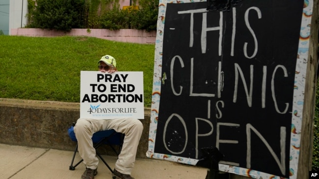 FILE - An abortion opponent sits behind a sign that advises the Women's Health Organization clinic is still open in Jackson, Mississippi, July 6, 2022. A report finds the number of monthly abortions in the U.S. is about the same after state bans started kicking in in 2022.