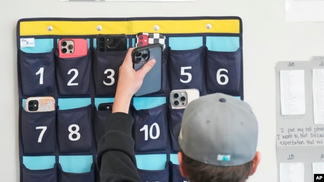A ninth grader places his cellphone into a phone holder as he enters class Feb. 23, 2024, in Delta, Utah. Each classroom has a cellphone storage unit that looks like an over-the-door shoe bag with three dozen smartphone-sized slots.
