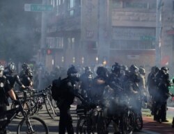 FILE - Seattle bicycle police officers wear gas masks and carry weapons as smoke rises and they clash with protesters, July 25, 2020.