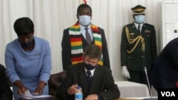 Andrew Pascoe (seated), the president of the Commercial Farmers Union of Zimbabwe, signs the agreement while President Emmerson Mnangagwa (with scarf) waits for his turn, at the State House in Zimbabwe, July 29, 2020. (Columbus Mavhunga/VOA) 