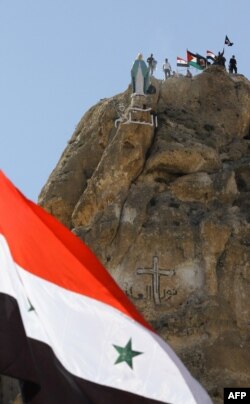 A Syrian national flag flutters in front of members of the Syrian National Defense Forces (NDF) standing behind a statue of Virgin Mary perched on the cliffs overlooking the ancient Christian town of Maalula, June 13, 2015.