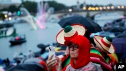 Una espectadora toma una foto con su celular mientras los deportistas pasan en barcos por el río Sena durante la ceremonia de inauguración de los Juegos Olímpicos de 2024 en París, Francia, el viernes 26 de julio de 2024. (Stefan Wermuth/Pool Photo víaAP).