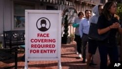 People without masks walk past a sign requesting customers to wear masks in Laguna Beach, Calif., May 17, 2021. 