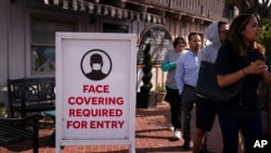 People without masks walk past a sign requesting customers to wear masks in Laguna Beach, Calif., May 17, 2021. 