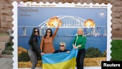 Gratë ukrainase pozojnë përpara një vepre arti në Kiev që pasqyron zjarrin në urën e Krimesë