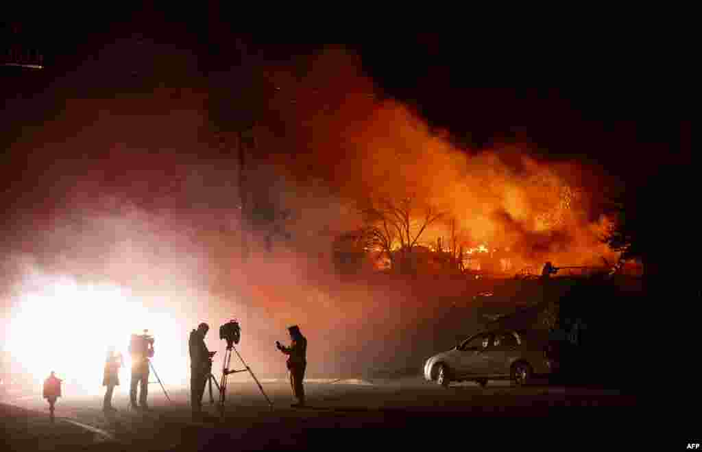 Media members report on a burning home as a firefighter douses flames (R) during the Hillside Fire in the North Park neighborhood of San Bernardino, California.