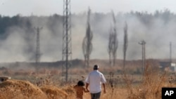 People walk in Akcakale, Sanliurfa province, southeastern Turkey, as in the background, Turkish forces artillery pieces fire targets towards Syria, Oct. 11, 2019.