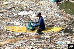 An environmental activist sits on a surf board as he picks up trash from a garbage-strewn river during a river clean up in Pecatu, Bali, Indonesia on Friday, March 22, 2024. (AP Photo/Firdia Lisnawati)