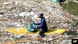 Seorang aktivis lingkungan duduk di atas papan selancar sambil memungut sampah dari sungai yang berserakan sampah saat bersih-bersih sungai di Pecatu, Bali, Jumat, 22 Maret 2024. (Foto: AP)