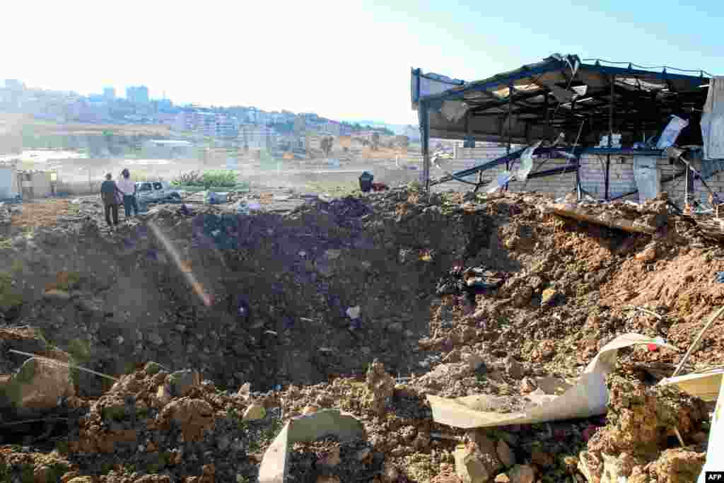 People stand by an impact crater next to a destroyed warehouse at the site of an Israeli airstrike in Jiyeh along the highway linking Beirut to the southern city of Sidon.