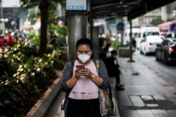 FILE - A woman, wearing a face mask, uses her mobile phone while waiting at a bus stop in Bangkok, Thailand, Jan. 30, 2019.