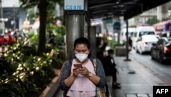 FILE - A woman, wearing a face mask, uses her mobile phone while waiting at a bus stop in Bangkok, Thailand, Jan. 30, 2019.