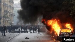 A car burns as protesters are seen during a rally against Expo 2015 in Milan, Italy, May 1, 2015. 
