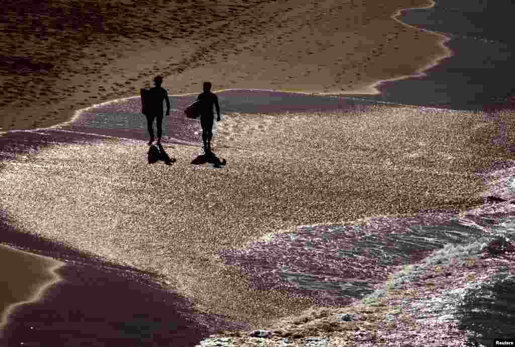 Dua orang peselancar berjalan di sepanjang pantai pada hari yang cerah di Pantai Bondi, Sydney, Australia.