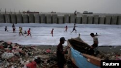 FILE - Fishermen repair their wooden boat as children play soccer near a new construction of a concrete sea wall in Cilincing district of Jakarta, Indonesia, Aug. 22, 2017.