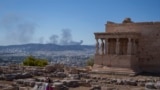 FILE - A woman poses for a photo in front Caryatid statues that prop up the porch of the 5th century B.C. Erechtheion temple, as at the background is seen smoke from a fire , during a hot day at Acropolis hill, in Athens, Tuesday, July 16, 2024. (AP Photo/Petros Giannakouris)