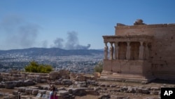 FILE - A woman poses for a photo in front Caryatid statues that prop up the porch of the 5th century B.C. Erechtheion temple, as at the background is seen smoke from a fire , during a hot day at Acropolis hill, in Athens, Tuesday, July 16, 2024. (AP Photo/Petros Giannakouris)