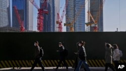 FILE - People wearing face masks walk by construction cranes near office buildings at the central business district in Beijing on March 15, 2023. 