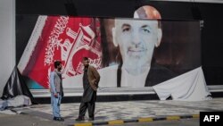 FILE - Afghan men stand next to a torn poster of deposed Afghan President Ashraf Ghani at Kabul airport, Aug. 16, 2021.