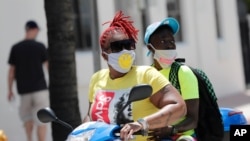 People wearing protective face masks ride a scooter down Ocean Drive amid the coronavirus pandemic, July 12, 2020, in Miami Beach, Fla.