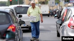 FILE - An Italian man walks between cars as he holds a banner reading "I'm a poor Italian, help me, thanks" in Rome, May 28, 2018. 