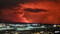 Houses in the village of Hafnarfjordur are seen as smoke billows in the distance and lava colors the night sky orange from a volcanic eruption on the Reykjanes peninsula, western Iceland on Dec. 18, 2023.