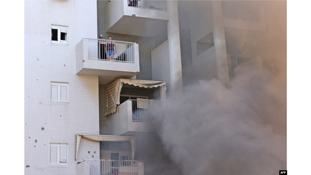 Seorang perempuan berdiri di balkon apartemennya sementara asap keluar dari tempat parkir gedung menyusul serangan roket dari Jalur Gaza di Kota Ashkelon, bagian selatan Israel, Sabtu, 7 Oktober 2023. (Foto: Ahmad Gharabli/AFP)