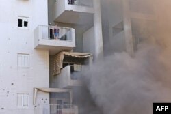 Seorang perempuan berdiri di balkon apartemennya sementara asap keluar dari tempat parkir gedung menyusul serangan roket dari Jalur Gaza di Kota Ashkelon, bagian selatan Israel, Sabtu, 7 Oktober 2023. (Foto: Ahmad Gharabli/AFP)