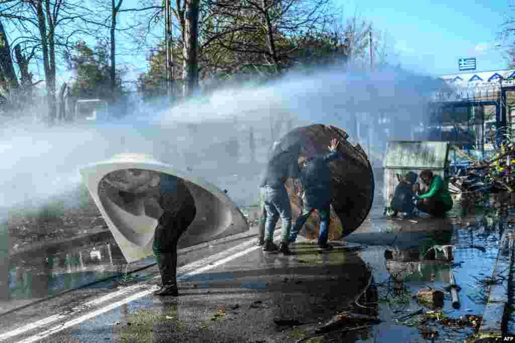 Migrants takes cover behind a trash container, a bathtub and a wooden board as Greek police use water cannons to block them trying to break fences in the Turkey-Greece border province of Edirne, March 7, 2020.