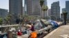 FILE - A homeless man sits at his street-side tent along Interstate 110 along downtown Los Angeles' skyline, May 10, 2018. Thousands of homeless people sleep on the streets of Los Angeles County. Southern California Public Radio station KPCC reports probl