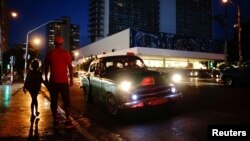 People wait on the sidewalk for transportation as a vintage U.S. car used as private taxi passes by in Havana, Cuba, Sept. 20, 2018.