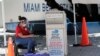 A poll worker wears personal protective equipment as she monitors a ballot drop box for mail-in ballots outside of a polling station during early voting, Aug. 7, 2020, in Miami Beach, Florida. 