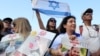 Orang-orang membawa poster yang memuat gambar sandera Israel yang baru dibebaskan, Yarden Bibas, serta kedua anaknya, Kfir dan Ariel, saat mereka menonton pembebasan sandera Israel di Jalur Gaza di layar di lapangan sandera di Tel Aviv pada 1 Februari 2025. (Foto: AFP)