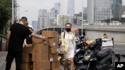 A woman wearing a mask walks past delivery workers moving boxes in Beijing on Aug. 20, 2020. U.S. and Chinese trade envoys discussed strengthening coordination of their government’s economic policies during a phone meeting Tuesday, Aug. 25, 2020,…