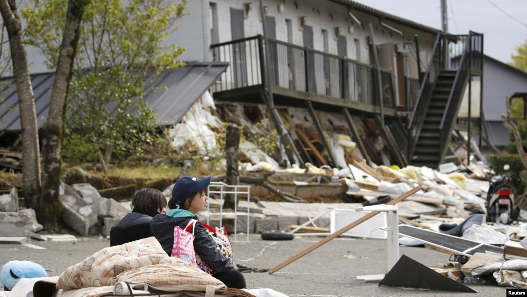 Mahasiswi beristirahat di depan gedung apartemen yang hancur akibat gempa bumi di kota Minamiaso di prefektur Kumamoto, Jepang selatan (16/4). 