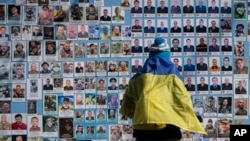 Seorang perempuan yang menyampirkan bendera Ukraina di pundaknya berdiri di samping Tembok Kenangan Para Pembela Ukraina yang tewas dalam Perang Rusia-Ukraina, pdi Kyiv, Ukraina, Selasa, 14 Maret 2023. (Foto: AP/Andrew Kravchenko)