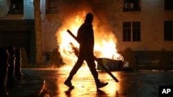 A man with a stick in hand walks past a burning police car not far from the Georgian parliament building in Tbilisi, Georgia, March 9, 2023. 